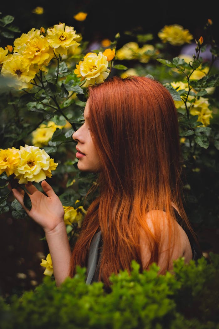 about-01 A woman with red hair enjoying the scent of blooming yellow roses in a garden.