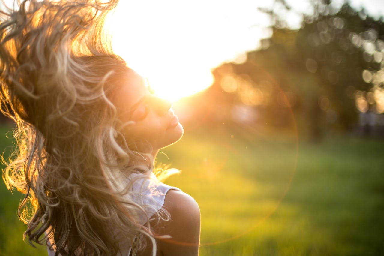 about-02 A young woman tosses her long, wavy blond hair in the sunlight during golden hour outdoors.